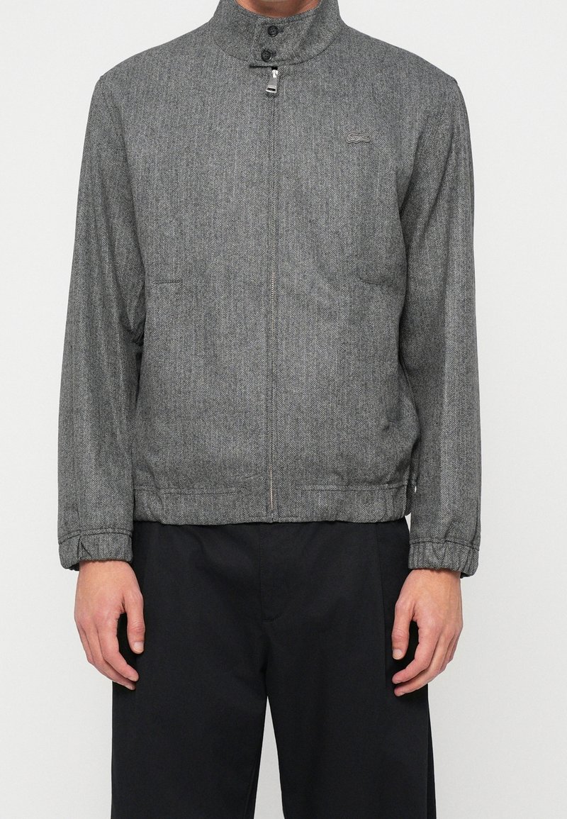 Homme portant une veste zippée grise en chevron avec col montant et pantalon noir, debout devant un fond blanc uni.