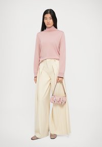 Maglione a collo alto rosa chiaro, pantaloni larghi color crema, che tiene in mano una piccola borsa rosa con accenti argentati, e scarpe a strisce multicolori.