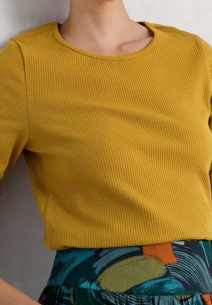 Frau trägt einen senfgelben, gerippten Oberteil und einen hoch taillierten Rock mit abstraktem grünem, blauem, orangem und schwarzem Muster.