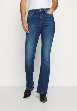 Vrouw draagt blauw denim jeans met middelhoge taille en licht uitlopende pijpen, gecombineerd met zwarte enkellaarsjes met puntige neus en een witte top die in de broek is gestopt.