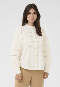 Camicia button-up color crema con un colletto alto, dettagli in pizzo intricati e maniche a sbuffo, realizzata in tessuto leggero.