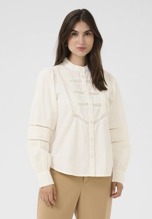 Chemise couleur crème à bouton, avec un col haut, des détails en dentelle élaborés et des manches bouffantes, fabriquée en tissu léger.