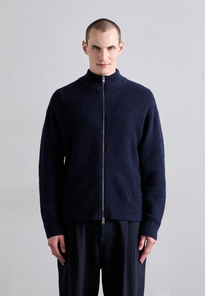 Chaqueta de cremallera azul marino con cuello alto, tejido texturizado y mangas largas. Presenta una cremallera de metal y un corte relajado.