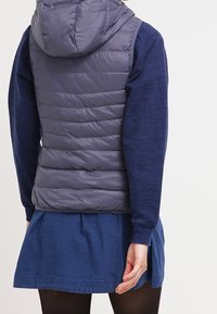 Graue Daunenweste mit Kapuze, gesteppter Design. Über einem blauen Pullover getragen, kombiniert mit einem blauen Rock. Das Outfit hat einen lässigen, geschichteten Look.