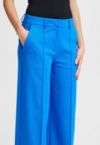 Platas kājas bikses spilgti zilā audumā, ar gludu tekstūru, priekšējām kabatām un plakanu jostasviju. Šūtas šuves piešķir struktūru.