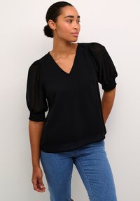 Zwarte blouse met een V-hals, opbollende doorschijnende mouwen en smock manchetten. De stof lijkt lichtgewicht met een gladde textuur, gecombineerd met blauwe jeans.