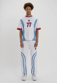 Balts sporta apģērbs, kas sastāv no īsām piedurknēm krekla ar zilām akcentiem un numuru 77, kopā ar atbilstošām biksēm ar zilām svītrām.