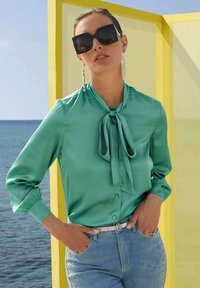 Eine grüne Satin-Bluse mit gebundenem Kragen, langen Ärmeln und Knopfverschluss, kombiniert mit hellblauen Jeans mit Verzierungen. Große schwarze Sonnenbrille.