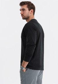 Mann in schwarzem Langarmshirt und grauen Nadelstreifenhosen, der seitlich mit den Händen in den Taschen vor einem schlichten Hintergrund steht.