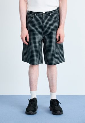 Black and teal checked shorts with a loose fit, featuring front pockets, a button closure, and subtle texture. Worn with black sneakers.