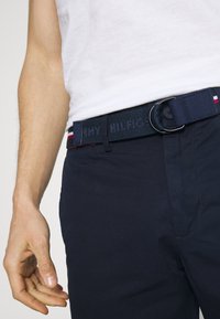 Marinblå bomullsbälte med en rund metallspänne, med broderat varumärke och röda och vita randiga detaljer i kanterna.