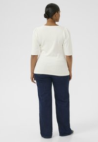 Kvinna med mörkt hår i en knut, klädd i en vit kortärmad topp och mörkblå jeans, står vänd bort från kameran mot en enfärgad bakgrund.