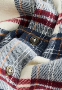 Ternet skjortestof med en blanding af marineblå, creme, rød og orange linjer. Indeholder en metal knap med indgraveret "SUPERDRY". Blød tekstur.