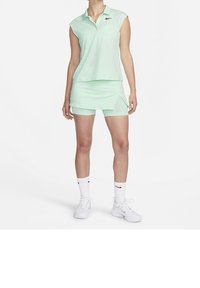 Femme portant un polo sans manches vert menthe, un skort assorti avec des shorts intégrés, des chaussettes blanches Nike et des baskets blanches Nike, debout sur un fond blanc.