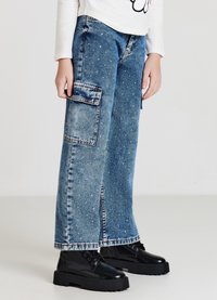 Pantalon cargo en denim bleu clair à coupe ample, avec poches avant et clous argentés dispersés. Associé à des bottes noires épaisses.