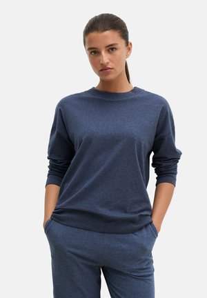 Frau mit dunklen zurückgebundenen Haaren, die ein passendes blaues langärmeliges Oberteil und eine Hose trägt, steht mit den Händen in den Taschen vor einem einfarbigen Hintergrund.