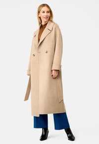 Beige wollen blend dubbelzijdig jasje met grote revers, zijzakken en een ontspannen pasvorm, gecombineerd met blauwe wijde jeans en zwarte enkellaarsjes.