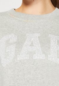 Jasnoszara bluza z dużym, błyszczącym napisem "GAP" z przodu. Materiał wydaje się miękki, ma okrągły dekolt oraz ściągacze przy rękawach.