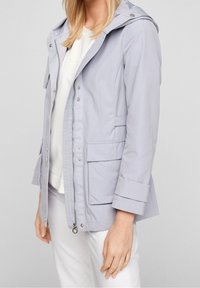 Veste imperméable gris clair avec capuche, fermeture éclair avant et deux poches latérales. Caractéristiques : poignets retroussés et tissu lisse et texturé.