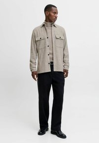 Grå herringbone skjortejakke med brystlommer og sorte knapper, båret over en beige turtleneck, parret med sorte bukser og sko.