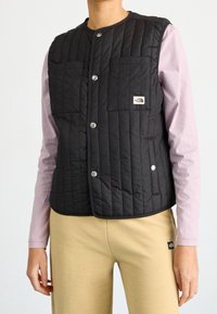 Chaleco negro acolchado con dos bolsillos frontales, cierre de botones a presión y pequeña etiqueta de logo, llevado sobre una camiseta de manga larga rosa claro.