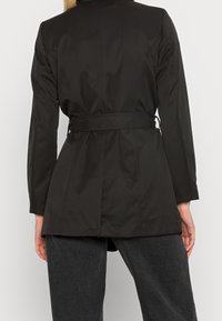 Svart trenchcoat med en figursydd silhuett, långa ärmar och ett bälte i midjan. Slät, matt tyg med rak ryggskärning och sidofickor.