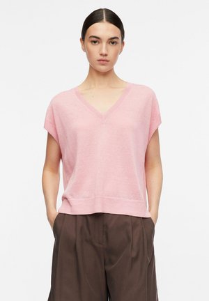 Frau trägt einen rosa V-Ausschnitt kurzärmligen Pullover und lockere braune Hose, steht mit den Händen in den Taschen vor einem einfarbigen Hintergrund.