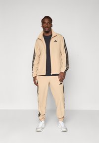 Survêtements beiges en matériau doux. La veste a un col montant et des rayures noires. Pantalons avec des rayures latérales noires, des poignets élastiques et un logo.