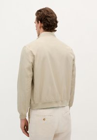 Chaqueta bomber beige claro con textura suave, puños y cuello acanalados; presenta una cintura ajustada y sutilezas en las costuras.