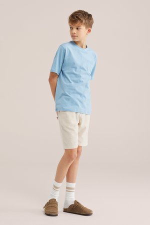 Băiat stând cu mâinile la spate purtând un tricou bleu deschis, pantaloni scurți bej, șosete albe cu dungi și pantofi maro slip-on.