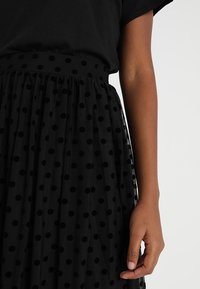 Musta pallokuvioinen tüllihame, jossa on kerätty vyötärö. Kangas on ohut ja läpikuultava, ja siellä on pyöreitä mustia samettipalloja, jotka tuovat pintaan tekstuuria.