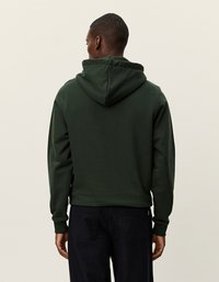 Zelena kapuca s gladko teksturo, rebrastimi manžetami in zaobljenim robom. Prikazana s hrbtne strani, čez temne hlače.