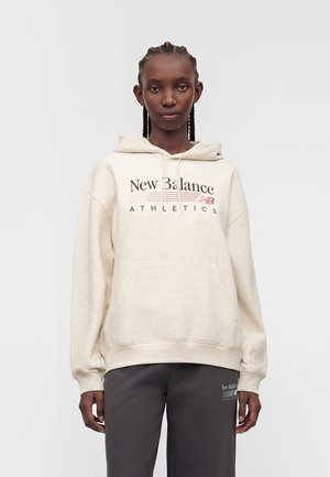 Γυναίκα μοντέλο με μπεζ φούτερ New Balance Athletics και σκούρο γκρι φόρμα, σε μονόχρωμο φόντο.