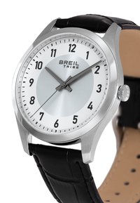 Montre analogique argentée avec cadran blanc, bracelet en cuir noir, chiffres arabes et un cercle texturé en argent au centre. Marque BRIEL TRIBE incluse.