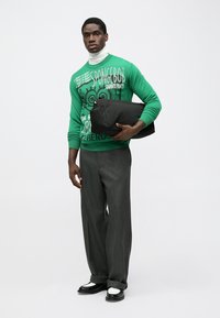 Green sweater featuring “SpongeBob SquarePants” graphics, paired with gray trousers and black shoes, holding a black bag.