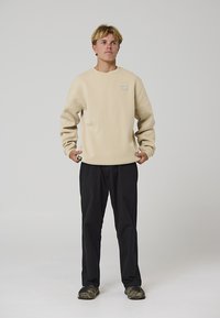 Sweat-shirt à col rond beige avec un petit patch de marque, associé à un pantalon noir et des baskets camouflage. Le tissu semble doux et texturé.