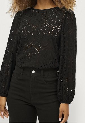 Nainen pukeutunut mustaan, pitkähihavaan pitsipaitaan ja korkeavyötäröisiin mustiin housuihin, esittäen osittain vartaloa ja käsiä.