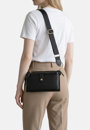 Donna con pantaloni beige e maglietta bianca che porta una borsa a tracolla in pelle nera testurizzata con ampia tracolla regolabile.