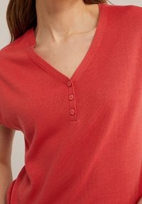 Koralowa czerwona bluzka z krótkim rękawem i dekoltem w kształcie litery V, z trzema małymi guzikami przy dekolcie oraz teksturowanym splotem materiału.