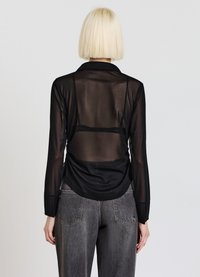 Blusa nera trasparente con maniche lunghe e colletto, caratterizzata da un design arricciato sul retro che si estende sotto la vita, abbinata a jeans grigi.