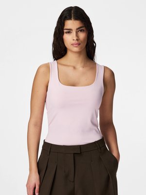 Νεαρή γυναίκα με μακριά σκούρα σγουρά μαλλιά που φοράει απαλό ροζ αμάνικο μπλουζάκι και σκούρο καφέ ψηλόμεσο παντελόνι με πιέτες.