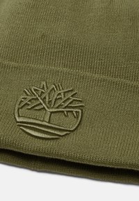 Olivově zelená čepice z měkké pleteniny, s vyraženým logem stromu v mírně světlejším odstínu, která předvádí žebrovanou texturu.