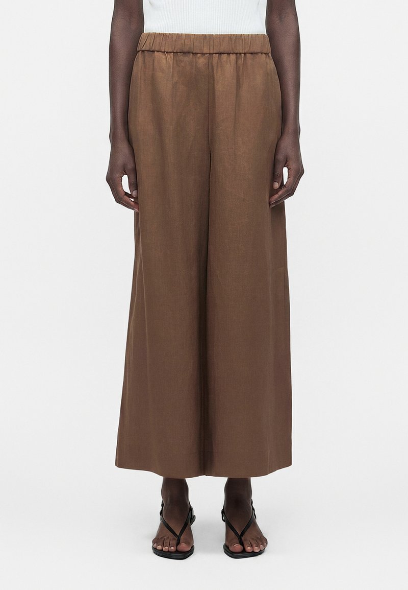 Καφέ φαρδιά cropped παντελόνια με ελαστική μέση, φορεμένα με μαύρα σανδάλια με λουράκια σε όρθιο άτομο.