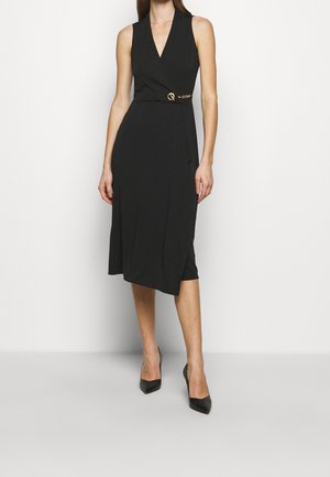 Femme portant une robe portefeuille midi noire sans manches avec un détail de boucle dorée et des escarpins noirs à bout pointu, debout sur un fond blanc.