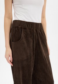 Brązowe spodnie sztruksowe z pionowymi prążkami, elastycznym pasem, boczną kieszenią i luźnym krojem, częściowo widoczne na jasnym tle.