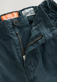 Tamno plave hlače s patentnim zatvaračem, koje imaju kopčanje na dugme. Vidljive oznake za Superdry i sportsku opremu. Glatka struktura tkanine.