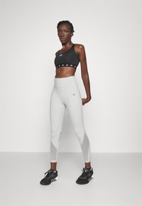 Sostén deportivo negro con logo blanco, leggings gris claro con cortes geométricos y zapatillas negras con rayas blancas. Textura suave en todo.