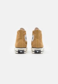 Zapatillas altas de lona marrón claro con suela de goma blanca, que presentan logotipos circulares en blanco y negro en el tobillo exterior y costuras blancas contrastantes.