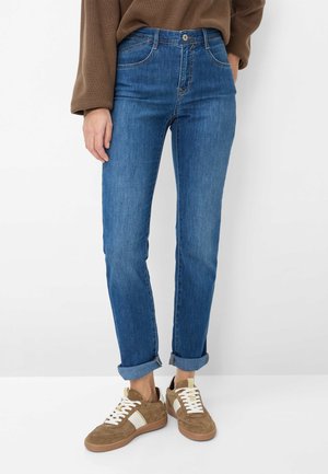 Person trägt hochgekrempelte blaue Jeans, braunen Pullover und braune Wildledersneaker mit weißen Schnürsenkeln, steht vor einem weißen Hintergrund.