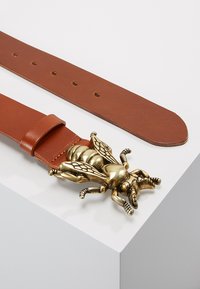 Brown leather belt featuring a large, detailed metallic bee-shaped buckle. The belt has multiple holes for size adjustment.